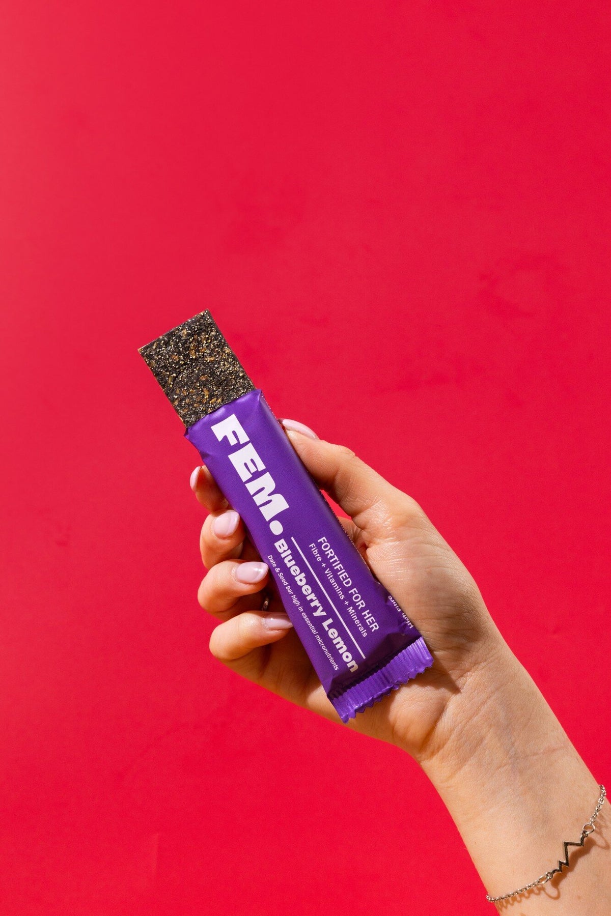 Hand holding an opened FEM. Blueberry Lemon bar against a red background